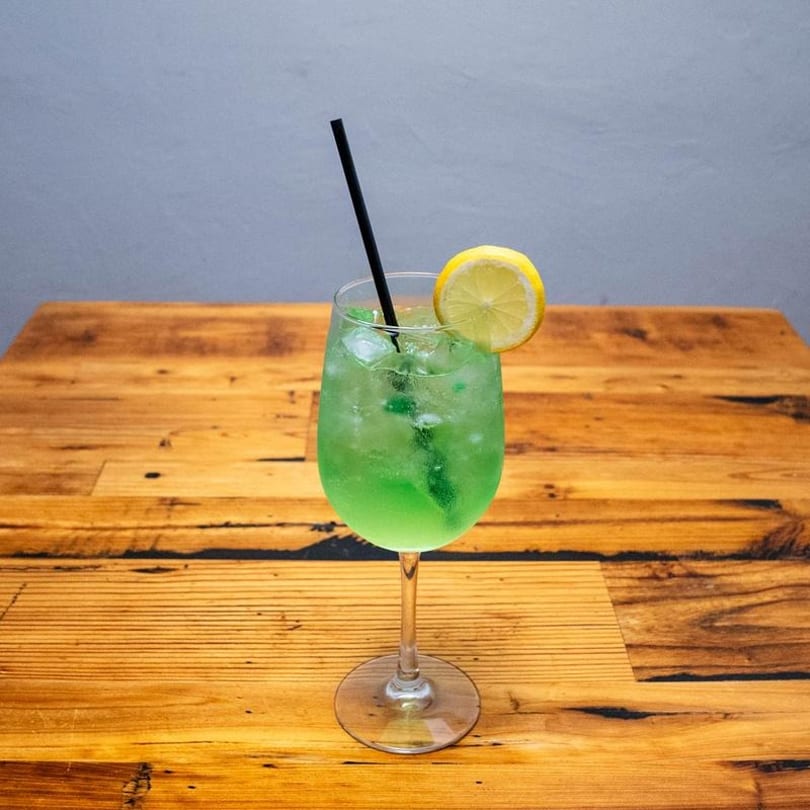 Green cocktail with ice, a black straw, and a lemon slice on a rustic wooden table.